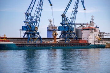 Fototapeta premium Industrial Harbor View of Blue Cranes Loading Grain or Sand into a Large Cargo Ship