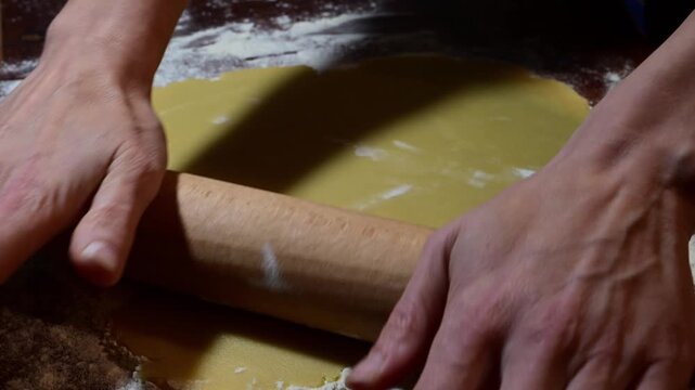 hands using a wooden rolling pin to flatten a sheet of yellow dough on a flour-dusted wooden surface.