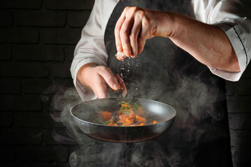 A chef in an apron prepares a dish, sautйing chopped vegetables in a frying pan. The cook adds salt as steam rises from the pan in a busy kitchen