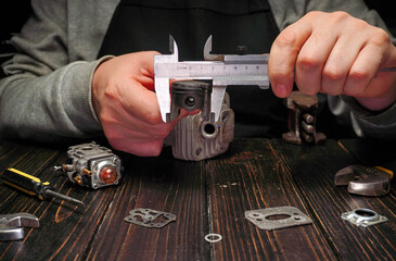 A person uses calipers to measure a small engine part on a wooden table. Several tools and parts are spread out, showing a workshop environment. Focus is on the precision of measurements