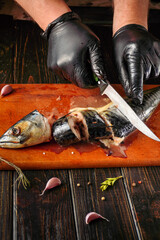 A person with gloves slices a fish on a wooden cutting board. Garlic and herbs are nearby. The kitchen has a rustic look with dark wooden surfaces © Віталій Б.