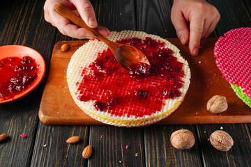 A person spreads red jam on a large wafer using a wooden spoon. Nearby, there are nuts and another wafer on a wooden cutting board. The scene is bright and simple
