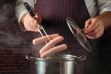 A chef prepares hot dogs by holding fork over a pot on the stove. The lid of the pot is being lifted while steam rises in a kitchen. The chef is focused on the task