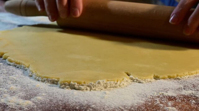 hands using a wooden rolling pin to flatten a sheet of yellow dough on a flour-dusted wooden surface.