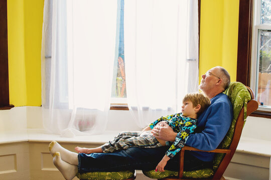 Side view of a young Boy sitting on his grandfather's lap on a chair by a bay window relaxing
