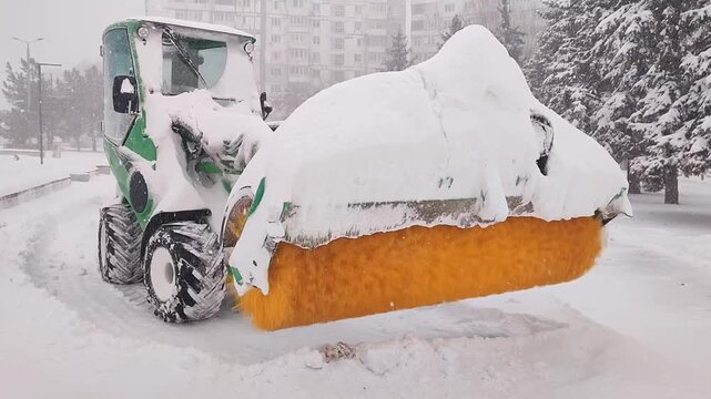 A mini tractor with a yellow shaft clears snow from park paths. Municipal equipment operates in challenging weather conditions.