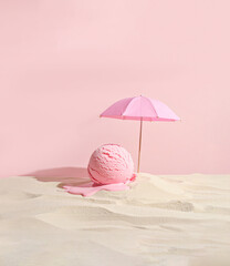 Conceptual layout of summer holiday and relaxation. The image depicts a scoop of pink, strawberry ice cream, melting on a patch of sand under a miniature pink beach umbrella against a pink background
