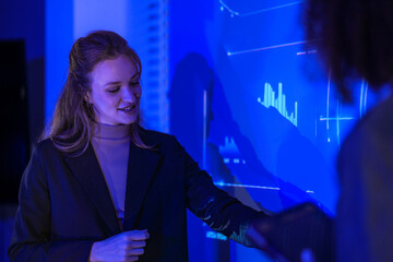 Female data scientist analyzing digital statistics on interface. Tech expert explaining software performance graph to team in modern blue control room.