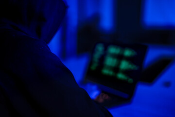 Mysterious hacker in black hoodie stealing confidential data on laptop. Cyber criminal coding dangerous malware virus to attack network system in dark blue room.