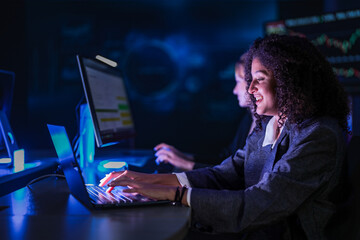 Dedicated business team working late on corporate project. Professional women analyzing digital performance data on screens in modern dark blue office at night.
