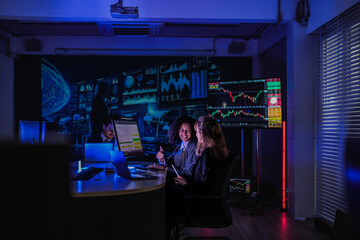Professional female traders analyzing stock market trends. Business team monitoring financial investment growth graphs on computer screens in modern night office.