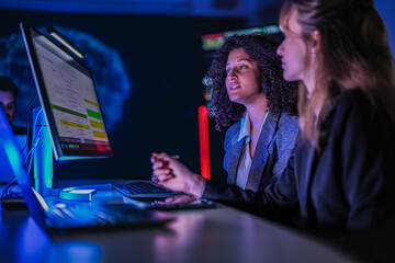 Cyber security analysts monitoring network data on digital display. Female tech experts discussing system defense strategy in dark control room center at night.