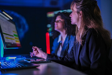 Dedicated business team working late on corporate project. Professional women analyzing digital performance data on screens in modern dark blue office at night.