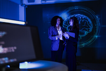 Businesswomen discussing financial strategy next to stock market screen. Professional team analyzing smart economy trends and AI data prediction in modern night office.