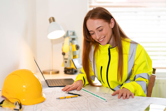 Young female professional builder in safety vest reviewing construction plans at workspace with laptop and yellow hard hat