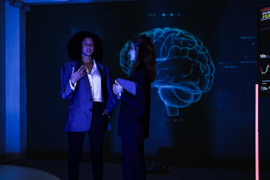 Businesswomen discussing financial strategy next to stock market screen. Professional team analyzing smart economy trends and AI data prediction in modern night office. - Powered by Adobe