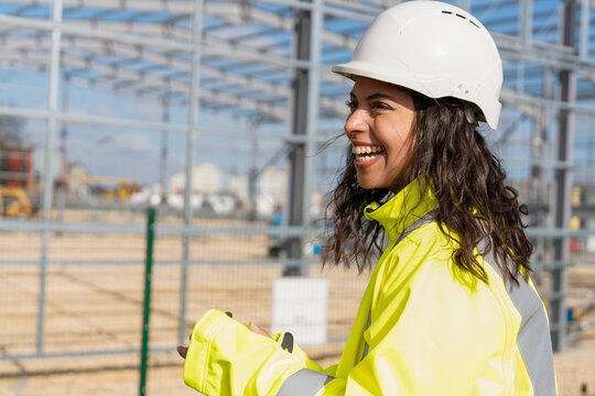 Happy smiling femail builder in hard hat and hi-viz fluorescent safety jacket in front of  steel frame structure in progress at construction site