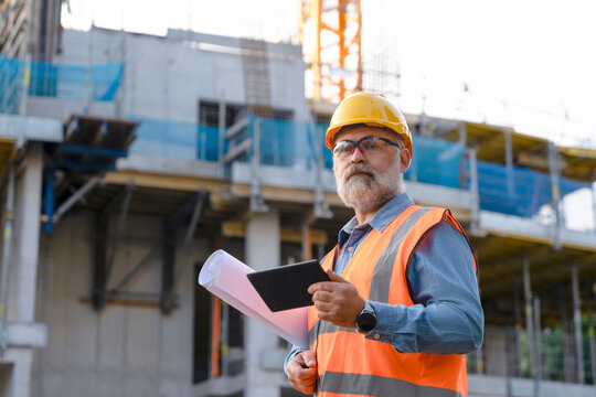 A construction supervisor in a safety vest and hard hat is observing the site, holding plans while overseeing building activity amidst cranes and construction workers in an urban area