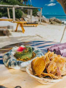 Fresh guacamole served with crispy root vegetable chips on a wooden table overlooking a sandy beach in Tulum, Mexico.