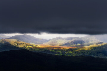 A picturesque view in Killarney National Park, Ireland. Sunlight breaks through a dark, stormy sky, illuminating the rolling green hills and valley below in a breathtaking scene.