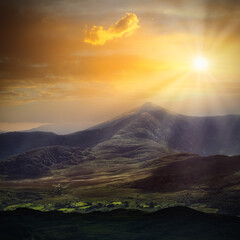 Golden sunlight spills across rugged mountain peaks and forested slopes. A single cloud drifts near the glowing sun above the valley. Warm hues highlight the contours of the central mountain.