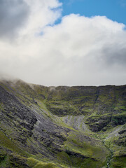 Clouds hover over a green and rocky mountainside, creating a scene of natural beauty and tranquility. A landscape of muted colors.