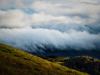 Green hills with grazing sheep are partially obscured by heavy fog and low clouds in the Highlands. The light shines on the hills creating a contrast with the approaching storm.
