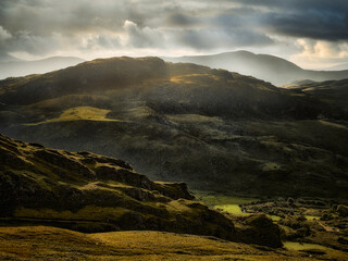 Green hills roll under a dramatic sky with rays of sunlight. The scene depicts a remote and natural landscape in National Park during daytime.