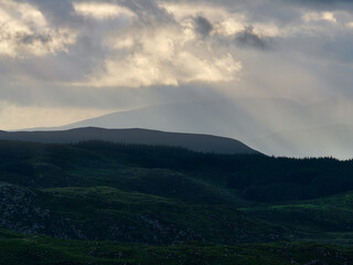 Sun rays break through grey clouds above rolling green hills and mountains of National Park. A dark forest borders a hill in the foreground.