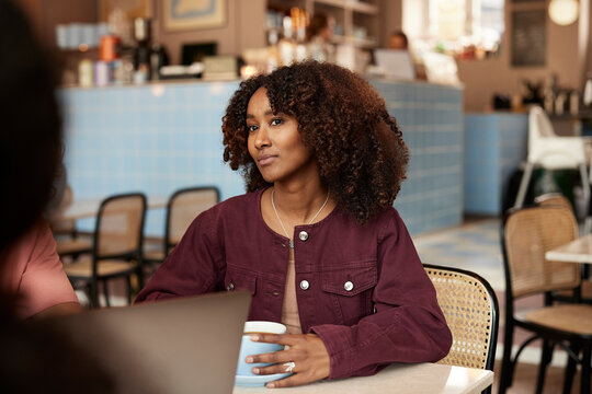 Woman in burgundy jacket sitting in a cozy cafe, holding a coffee mug and looking thoughtful