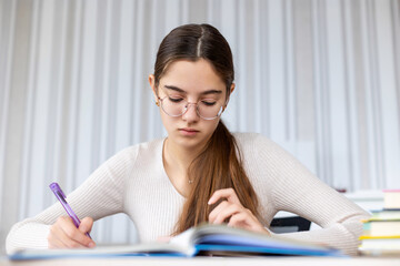 Focused student studying and taking notes indoors. A teen girl wearing glasses studies intently at a desk, flipping through a book and writing notes in a quiet room.