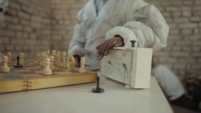Close up of chess game in progress with worn clock on table as person in dirty white coat taps timer after making move, with various white and black pieces arranged in tense strategic formation