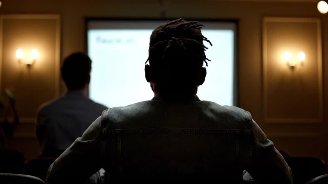 Attendee watches presentation on screen. Audience and viewer face presenter. Education training fits conference meeting. Student and listener observe attentively. Dim warm light auditorium atmosphere.