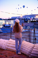 Obraz premium Relaxed woman in hat looks at hot air balloons floating in sky of Cappadocia. Traveler stands on rooftop, gazing at colorful balloons at dusk. Concepts: travel, summer vacation, impressions