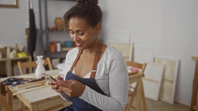 Woman wearing apron holding smartphone and smiling while checking messages at art studio worktable; creative calm.