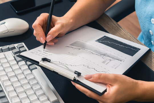 Analytics, finance. A woman's hand writing with a pen on a piece of paper with data and graphs. A keyboard, computer mouse, and smartphone on the table. Close-up. Top and side view. - Powered by Adobe