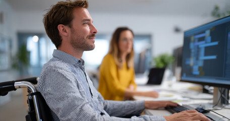 Young man in wheelchair working on computer in modern environment with female colleague in background