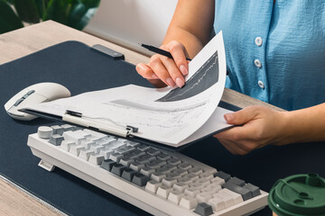 The manager analyzes and studies documents with data and graphs in a notebook. There is a keyboard and a computer mouse on the table. Close-up. Side view.