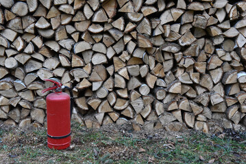 Red portable fire extinguisher standing on the grass next to a large stacked firewood pile