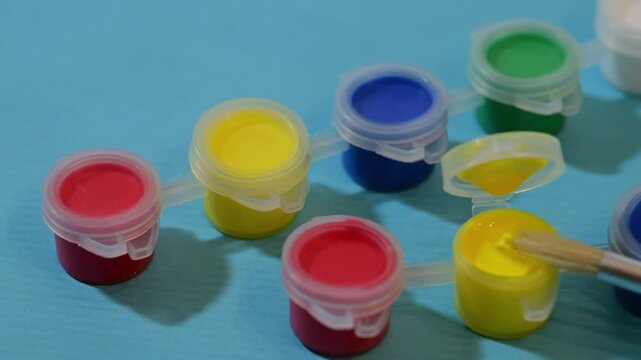 a paintbrush being dipped into a small pot of yellow paint within a colorful strip of plastic containers on a blue background.