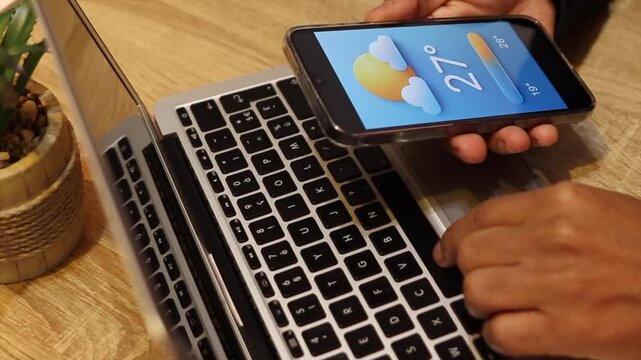Person checking the weather forecast on a smartphone while typing on a laptop computer. Business professional multitasking at a wooden office desk