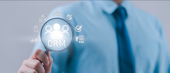 Businessman holding magnifying glass over CRM icon, representing customer relationship management, data analytics, sales strategy, and digital tools for improving client engagement and retention.