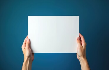 Human hands hold white paper sheet against blue background. Person shows empty blank paper with space for text message advertisement. Concept of presentation announcement or advertising.