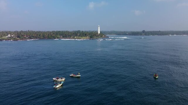 Aerial drone photo of Dondra Head Lighthouse in Sri Lanka with traditional fishing boats and fishermen in the foreground. Tall white lighthouse standing on the southernmost point of the island.
