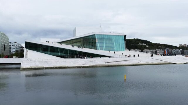 visitors walk across sloping white marble roof oslo opera house angular lines calm fjord waters city buildings hills appear norway expansive terrace surrounding 