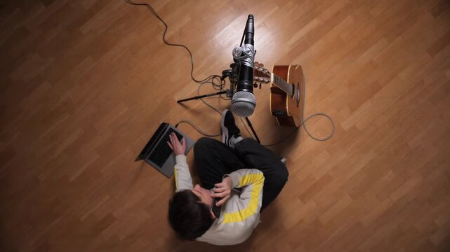 Musician preparing for an online live performance, typing on a laptop with a microphone and acoustic guitar set up on a wooden floor, connecting with an audience from a personal space