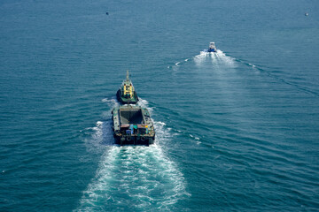 A tugboat pulls a cargo ship through calm waters close to the shore. The sky is clear, and the vessels create wakes as they move together. The scene shows activity on the water