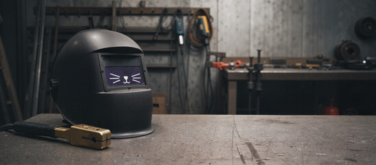 Welding helmet with cat face illustration sitting on metal workbench in industrial workshop setting