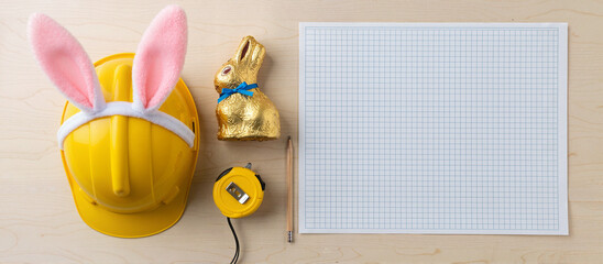Yellow hard hat with pink bunny ears and chocolate rabbit sitting on wooden desk next to graph paper and measuring tape