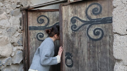 A brunette girl in a gray knitted sweater with openwork sleeves looks into an old wooden door surrounded by stone walls.